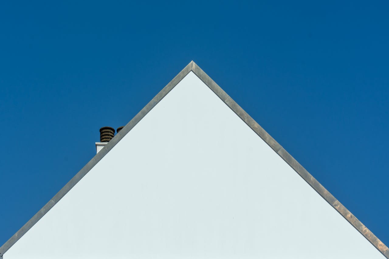 gallery-1 Triangle gable of a white house with chimney against a clear blue sky, highlighting minimalist architecture.